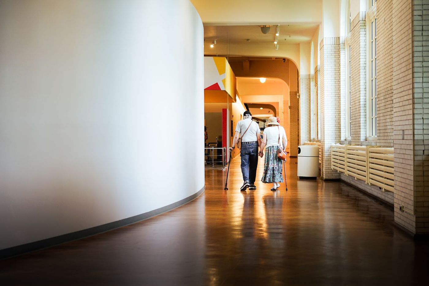 An older couple holding hands while walking away down a sunlit hallway, each using a cane