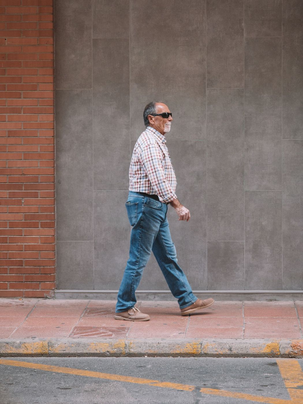 An older man in a plaid shirt and sunglasses walking in profile along a sidewalk in Valencia, Spain, mid stride