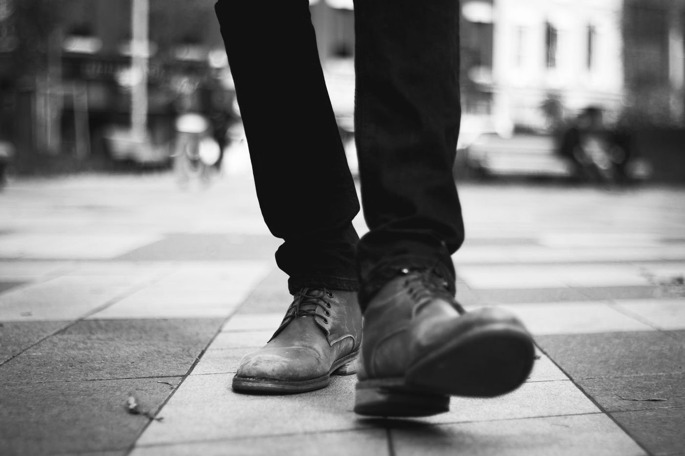 Black and white close up of boots mid stride on city pavement, one foot flat and one heel raised
