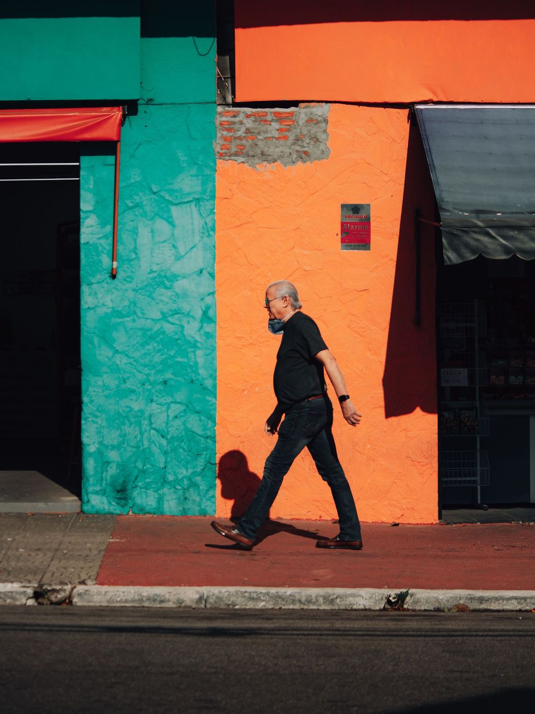 An older man walking in profile past a bright orange and teal wall, caught mid stride