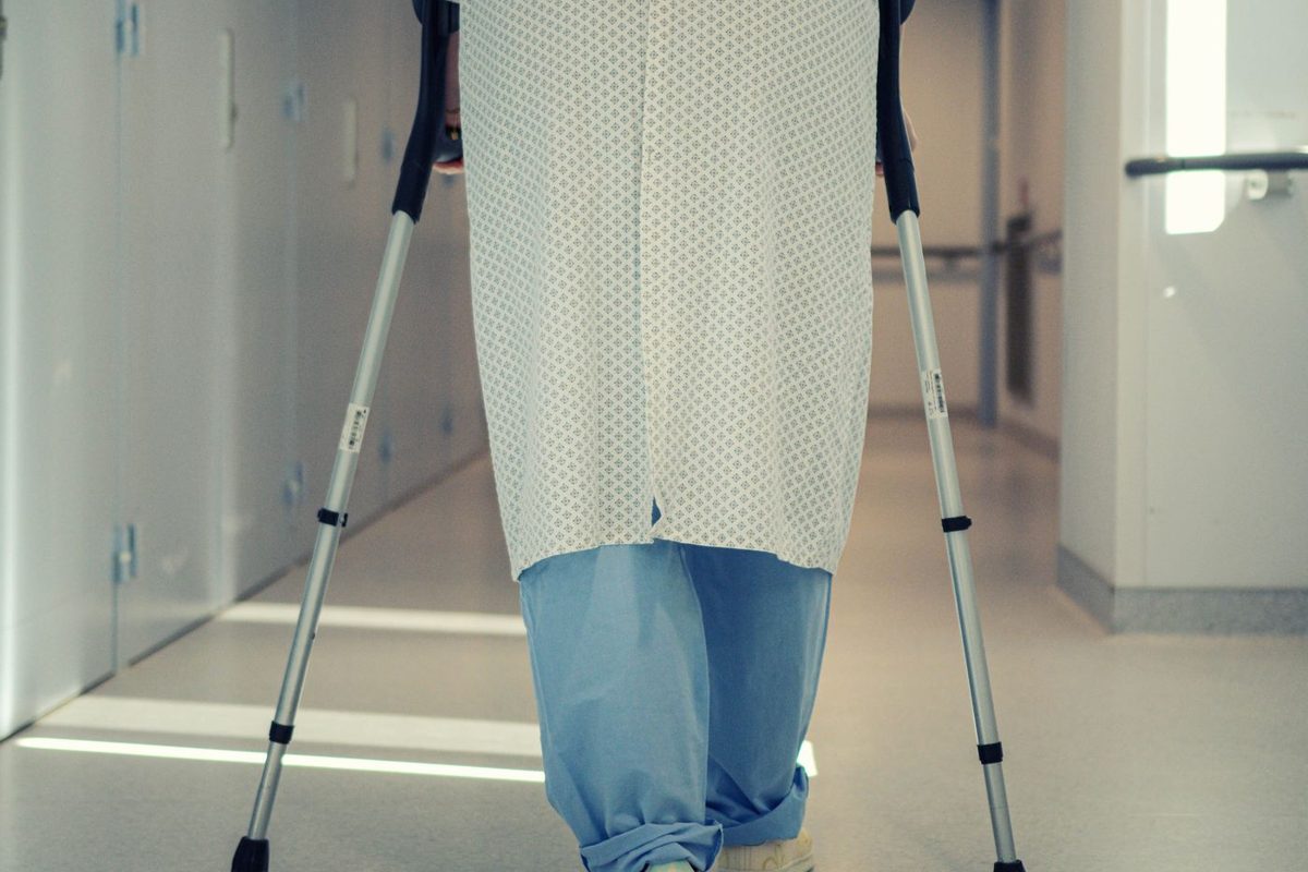 A patient in a hospital gown walking away down a corridor on crutches, seen from behind