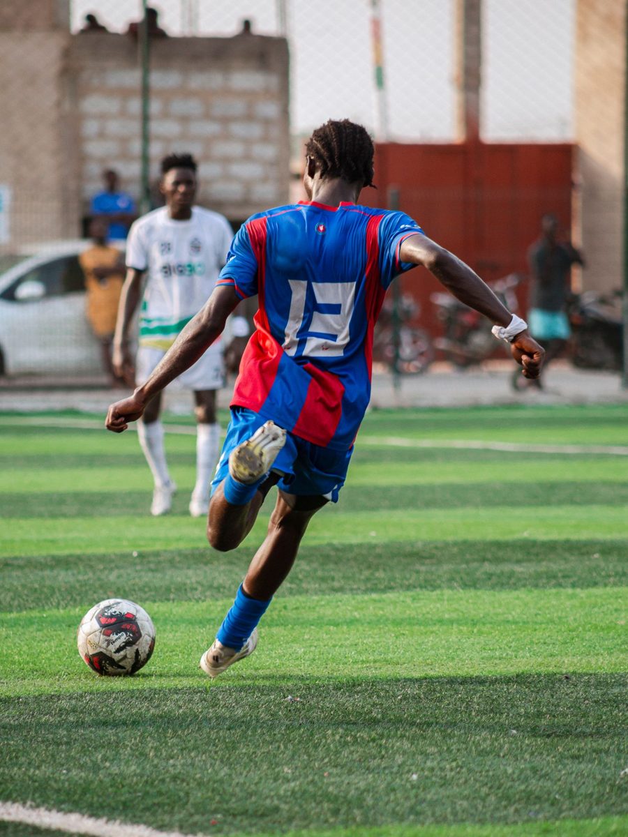 A young soccer player in a blue and red kit striking the ball on a turf field, seen from behind