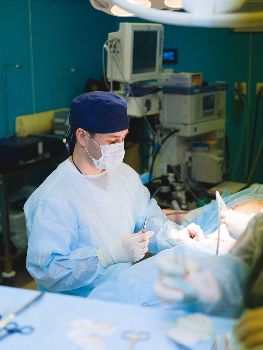 A surgeon in cap, mask, and gown suturing at an operating table under bright lights, the patient draped