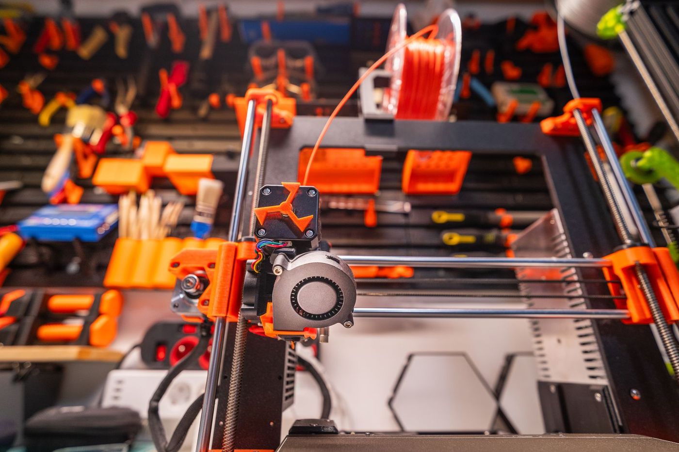 A 3D printer on a workshop bench with a pegboard of tools and a spool of orange filament behind it
