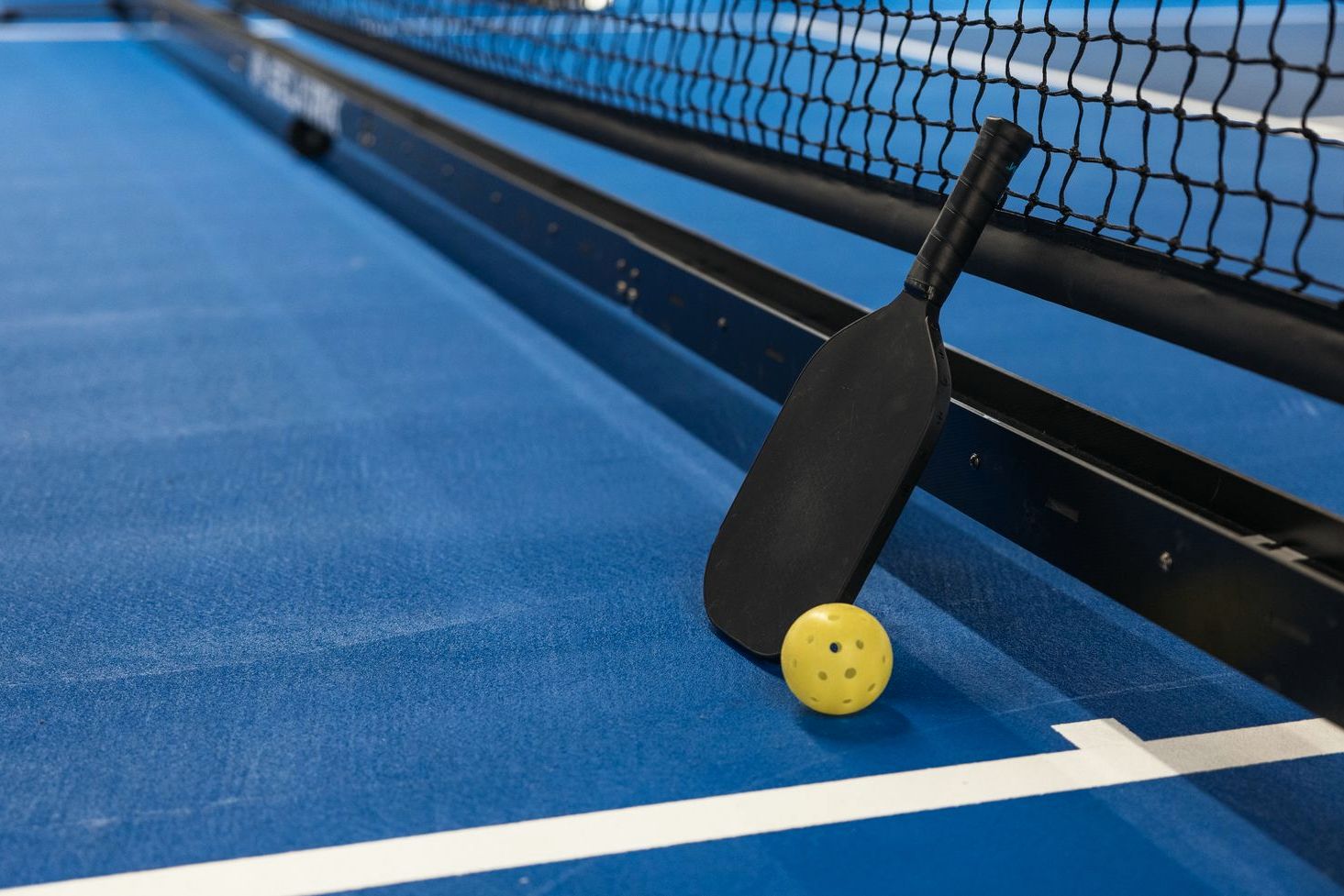 A pickleball paddle and yellow ball resting against the net on a blue court
