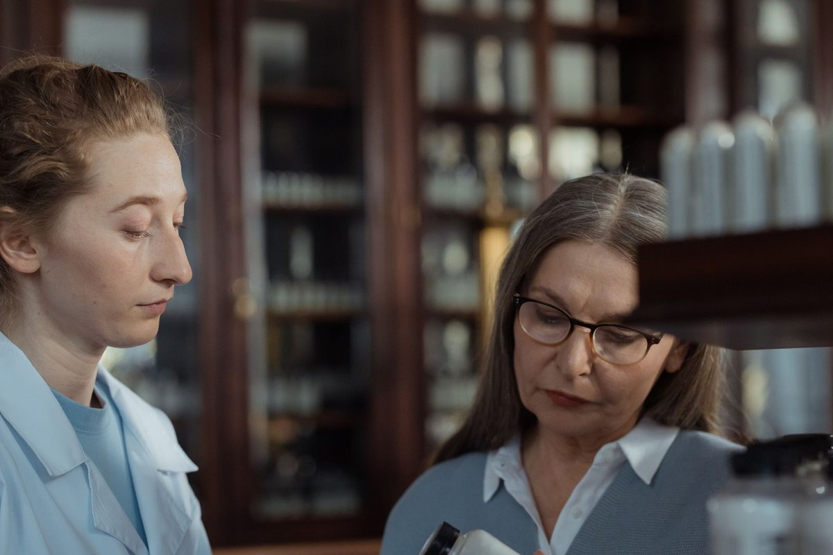 A pharmacist helping an older woman read a plain white medicine bottle at a pharmacy counter