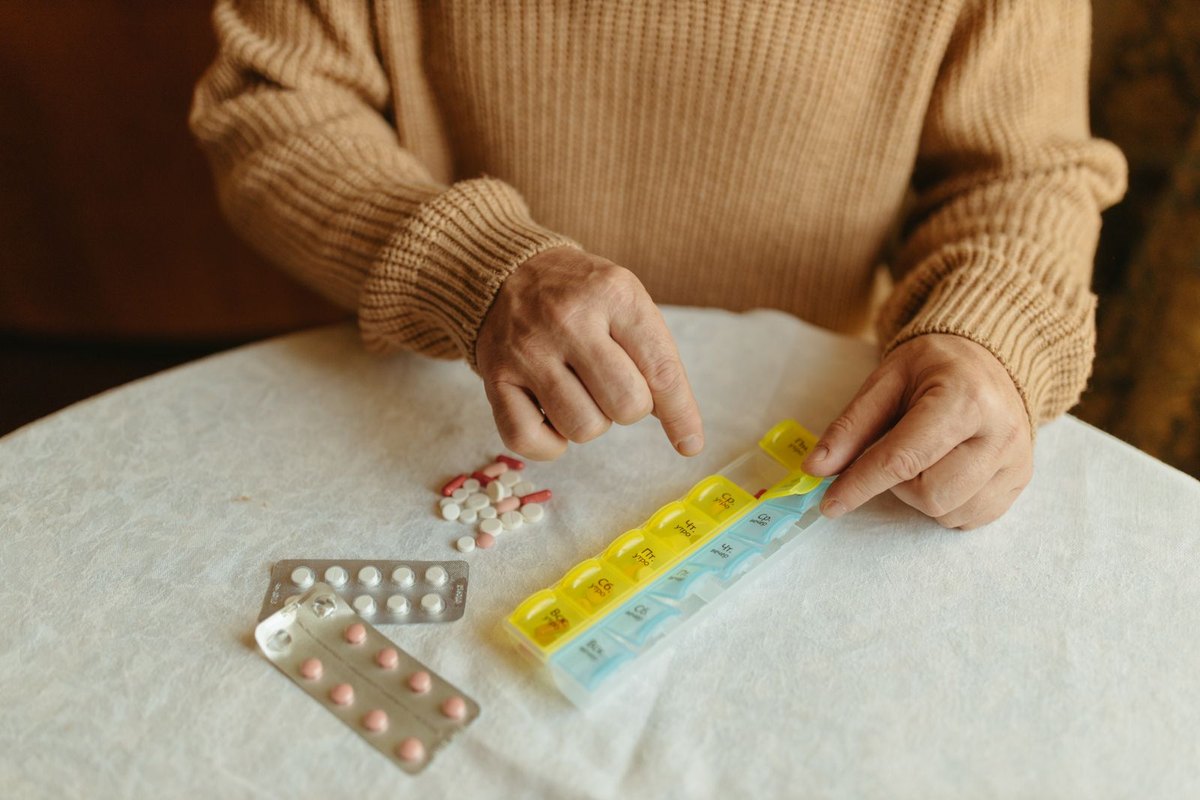 Hands sorting tablets and blister packs into a weekly pill organizer on a table