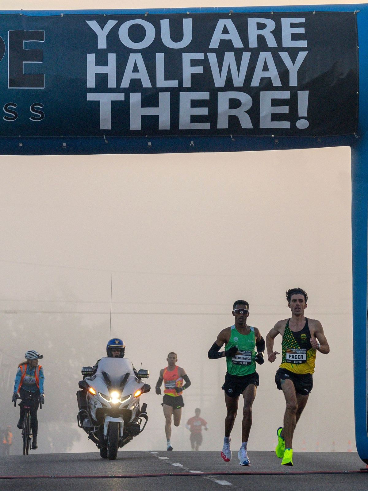 Lead runners passing under a banner that reads You Are Halfway There, with a motorcycle escort