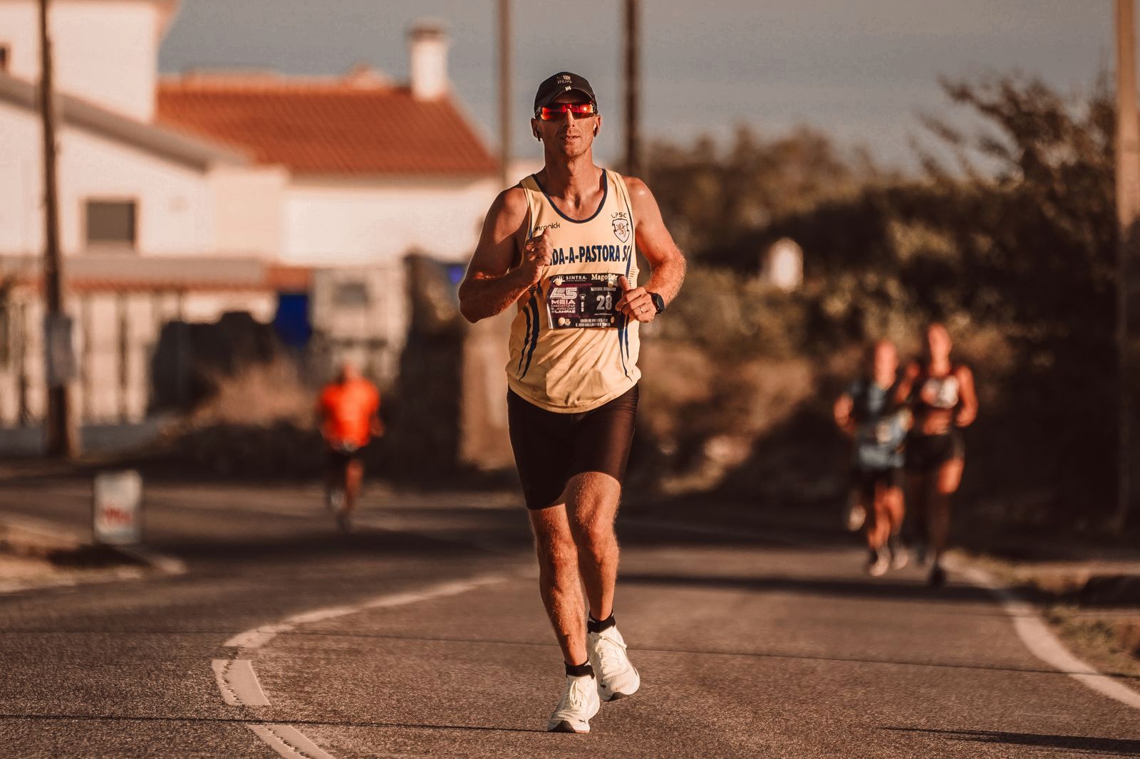 A lone runner in sunglasses and a race bib on a sunlit road, other runners blurred behind